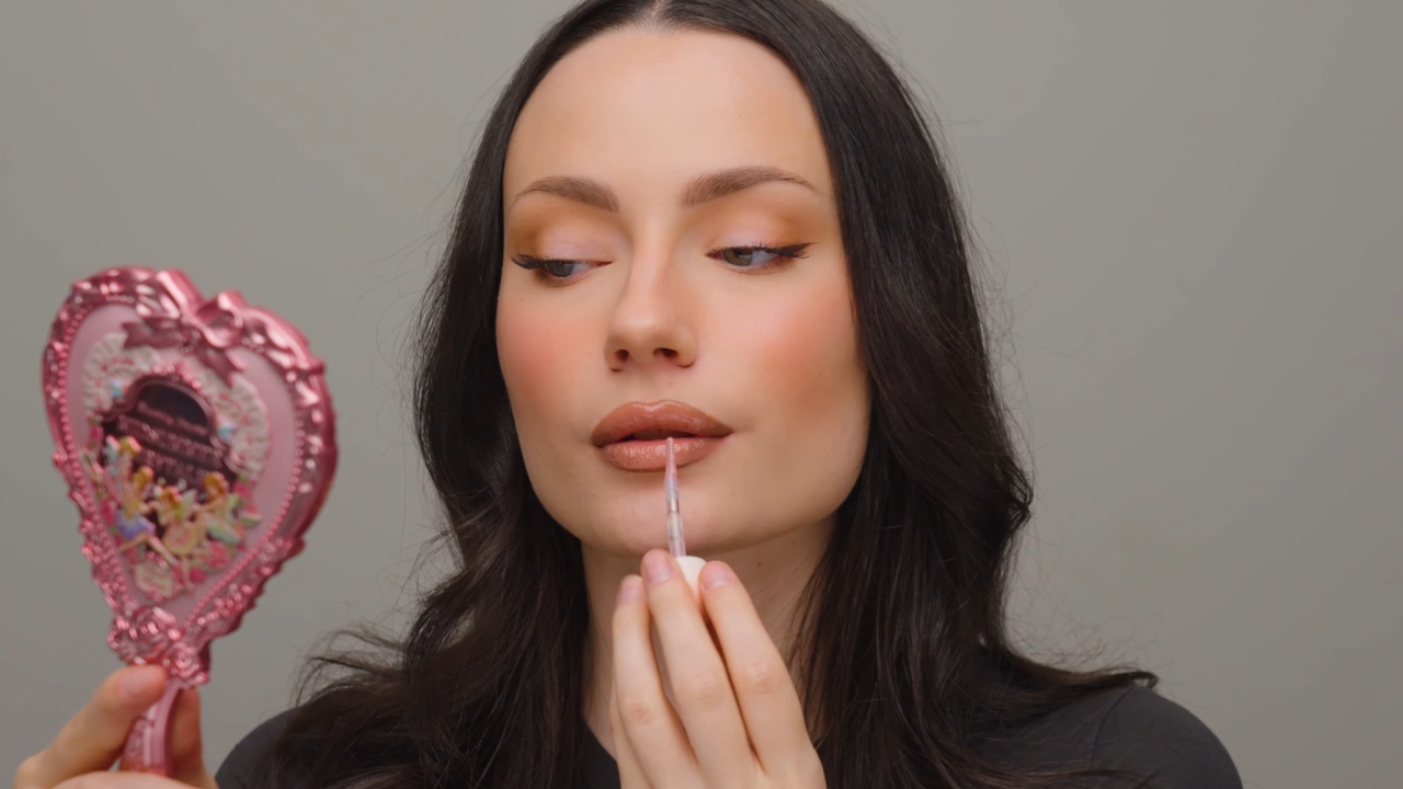 Žena nanosi nude sjaj na usne uz ogledalo, sa prirodnim tenom i mekim konturama usana